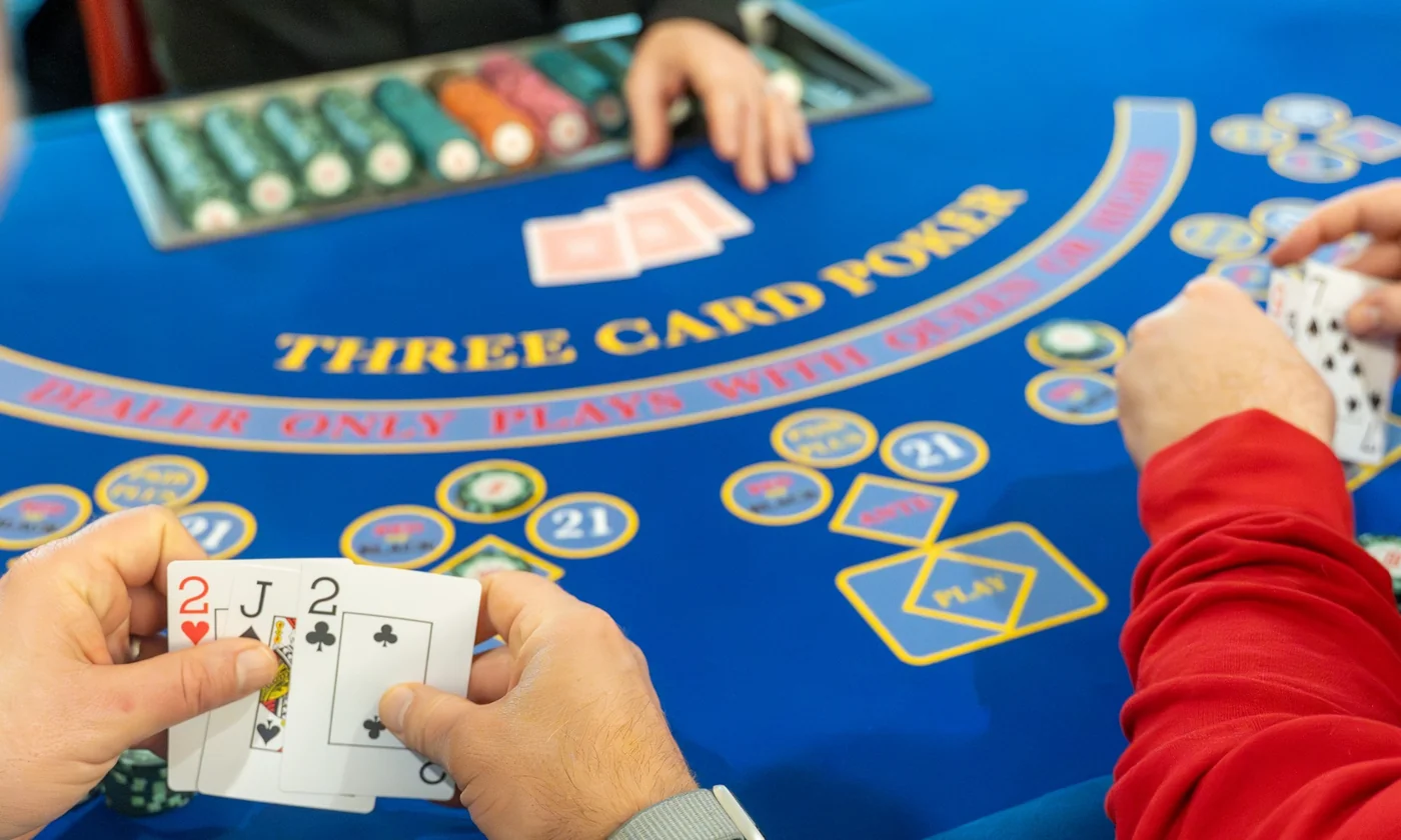 A blue Three Card Poker layout mid-play, a player holding cards over the Ante and Play diamonds with the chip tray behind